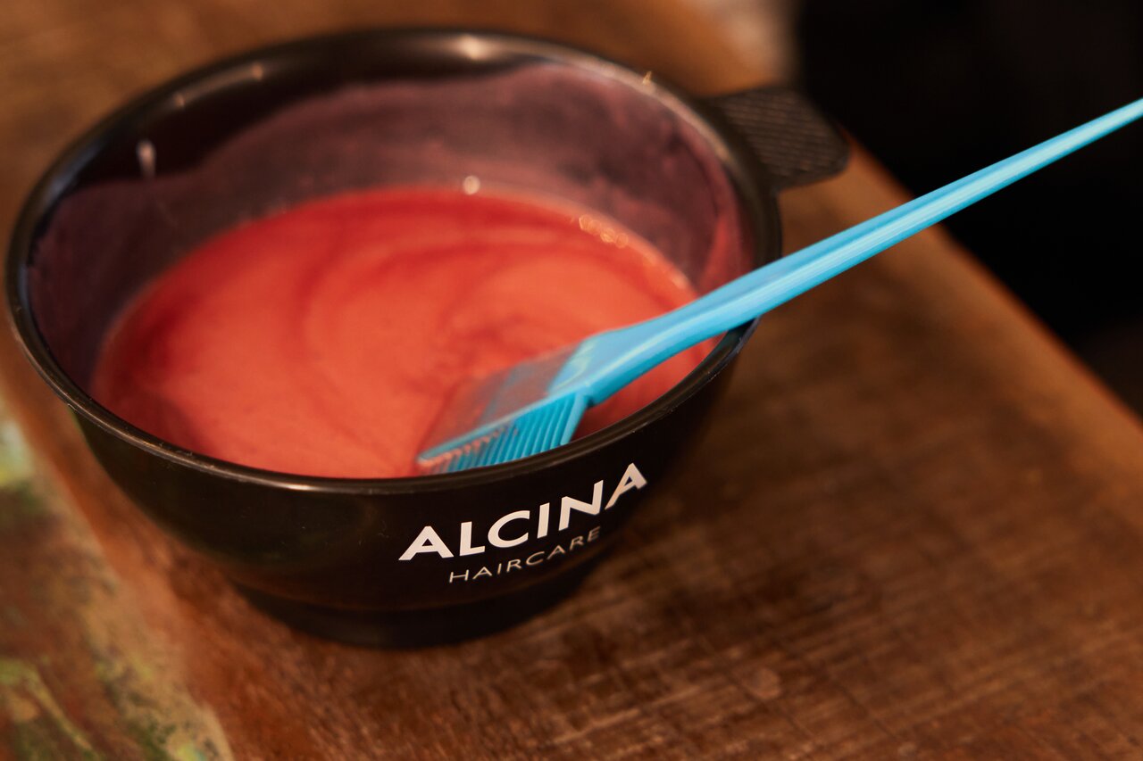 ALCINA bietet trendsichere Haarfarben für individuell und perfekt zusammengestellte Farbtöne. Nahaufnahme einer mit roter Haarfarbe gefüllte Farbschale, darin eingetaucht ein türkisfarbener Haarfärbepinsel.