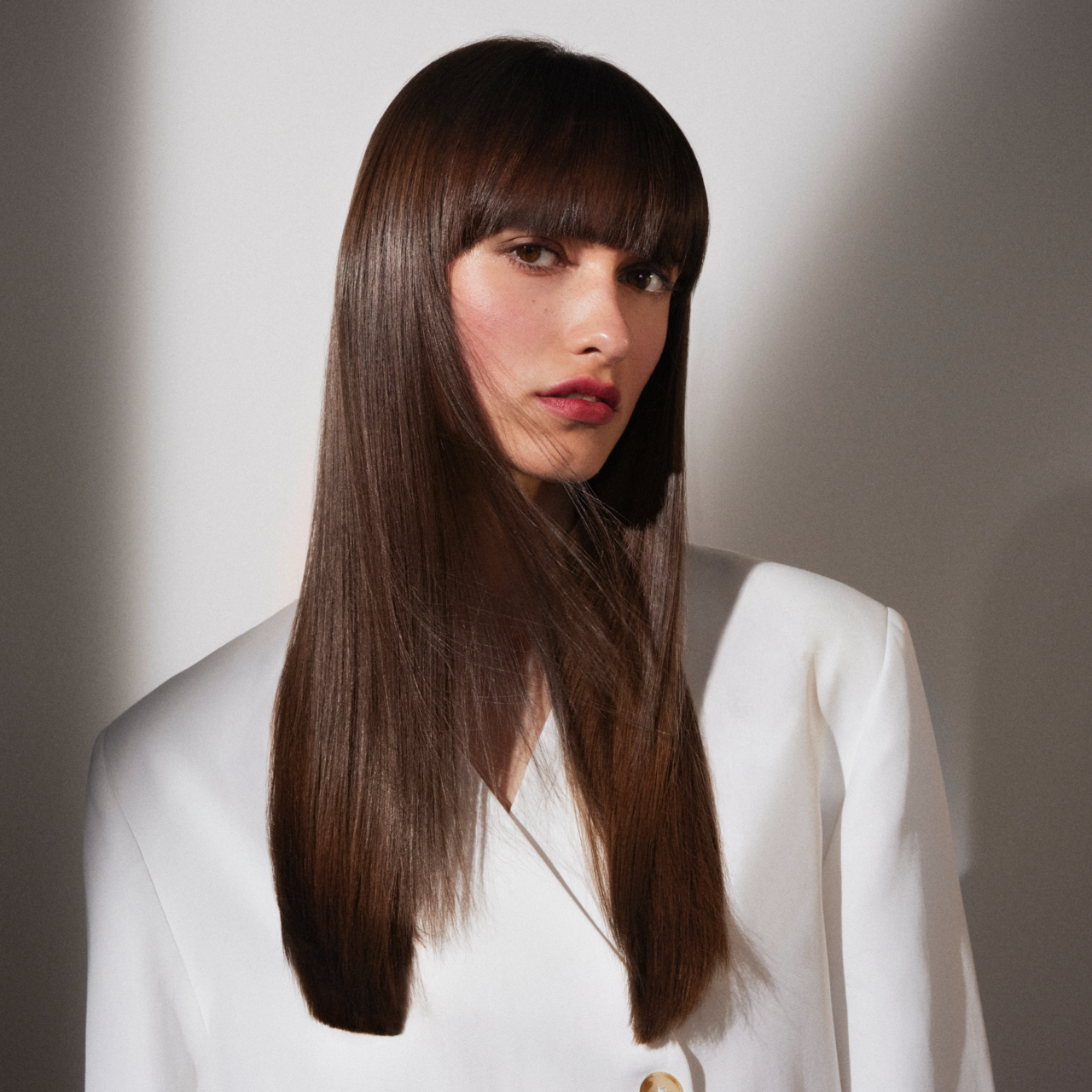 Frau mit langem, glänzend glattem dunkelbraunem Haar und geradem Pony trägt eine weiße Bluse und betont mit dezentem Make-up und roten Lippen einen eleganten, gepflegten Look.