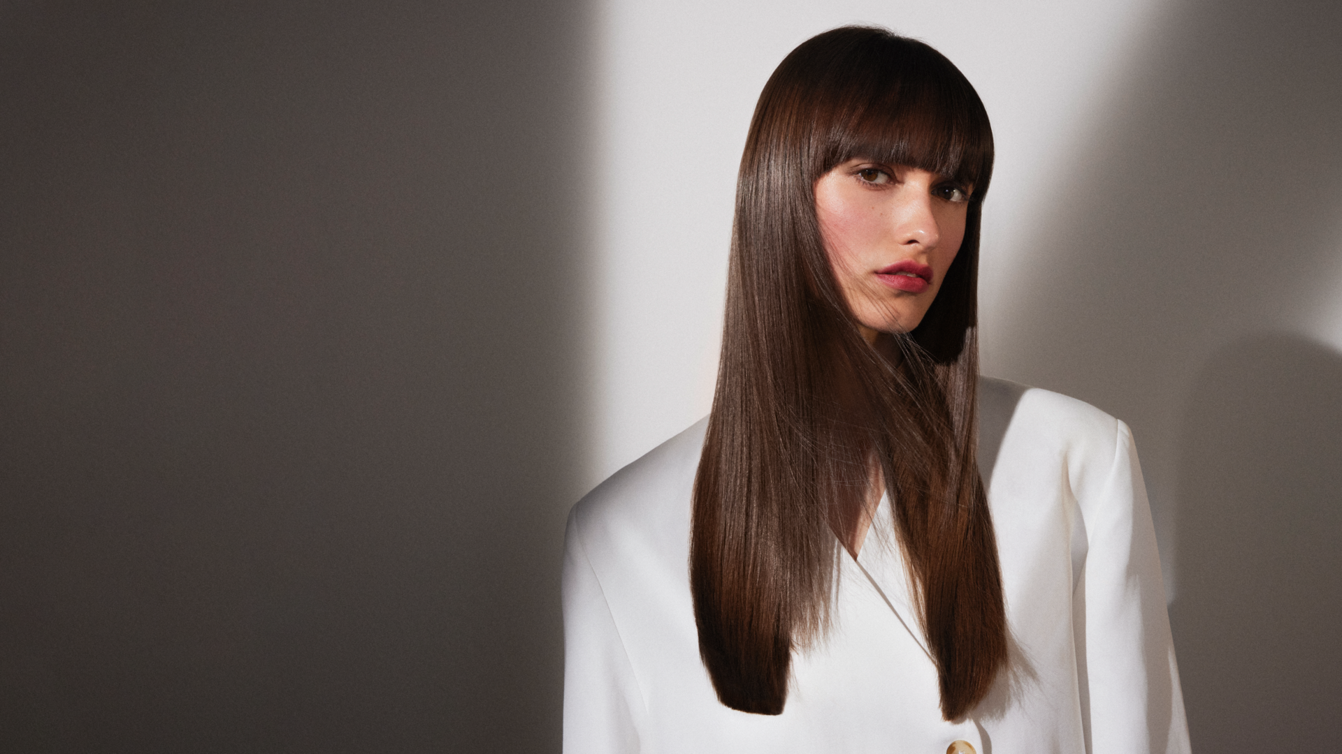 Person mit langem, glatten, glänzenden Haar in einem weißen Blazer, minimalistisch vor hell-dunklem Hintergrund inszeniert.