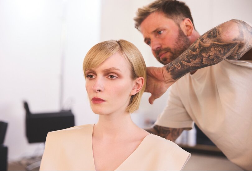 Eine Person mit kinnlangen blondem Haar sitzt, während eine andere Person, tätowiert, hinter ihr steht und ihre Haar stylt. Sie befinden sich in einem hellen, modernen Studio.