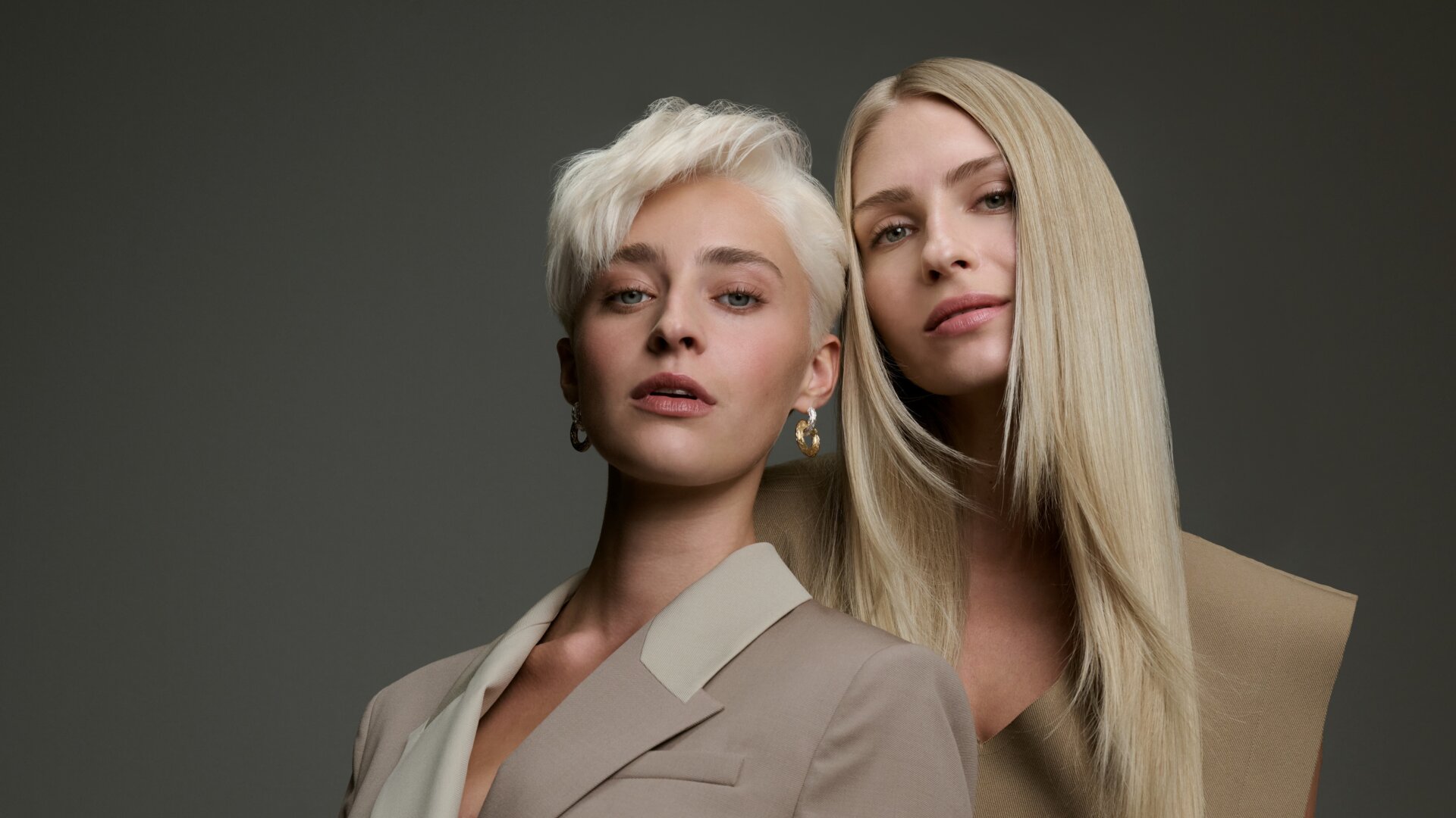 Zwei Models mit blonden Haarvarianten – ein kurzer platinblonder Pixie-Cut und glattes, langes Haar in warmem Blond – präsentieren vielfältige Blondtöne vor einem dunkelgrauen Hintergrund.
