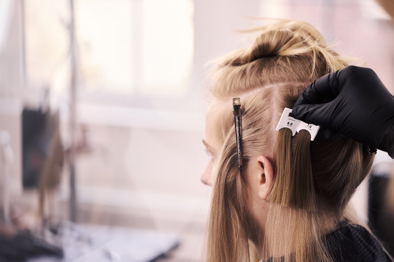 Friseur vergleicht mit einer Farbkarte den Naturton am Haaransatz einer blonden Kundin als Teil der professionellen ALCINA Blond-Farbberatung im Salon.