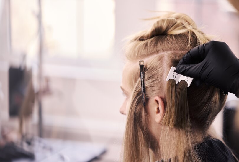 Friseur vergleicht mit einer Farbkarte den Naturton am Haaransatz einer blonden Kundin als Teil der professionellen ALCINA Blond-Farbberatung im Salon.