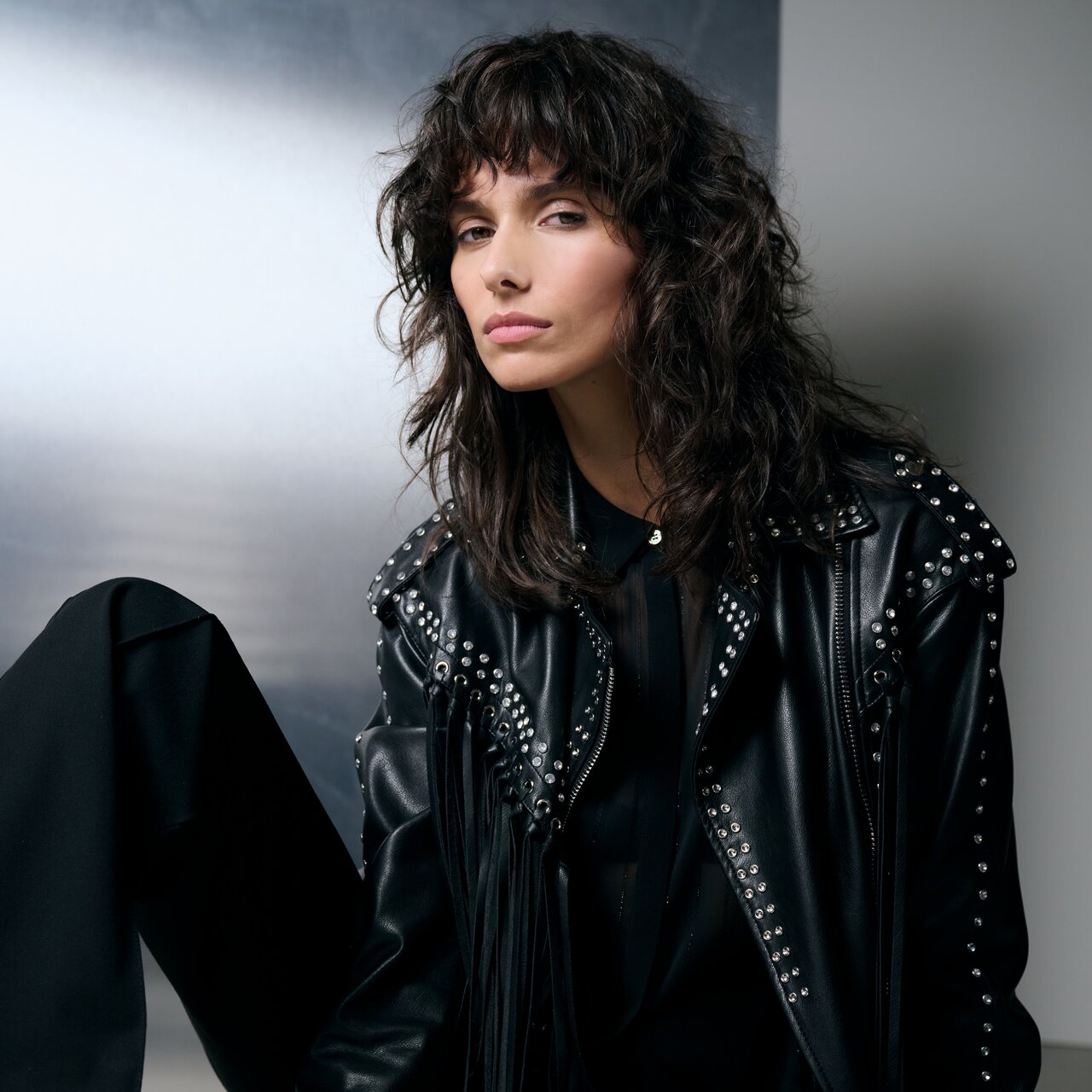 Junge Frau mit schulterlangem, wellig gestylten Haaren in einer schwarzen Lederjacke mit Nieten vor metallisch-grauem Hintergrund, die einen rockigen, edgy Look präsentiert.