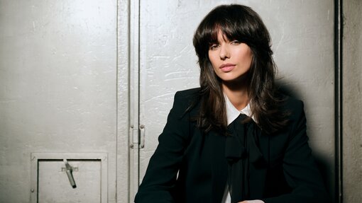 Person mit schulterlangem, voluminösem Haar im dunklen Hosenanzug und weißer Bluse mit Schleife, lässig vor einer metallischen Industrietür sitzend und einen eleganten Business-Look ausstrahlend.