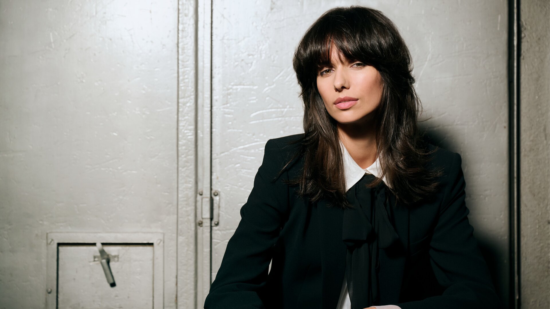 Person mit schulterlangem, voluminösem Haar im dunklen Hosenanzug und weißer Bluse mit Schleife, lässig vor einer metallischen Industrietür sitzend und einen eleganten Business-Look ausstrahlend.