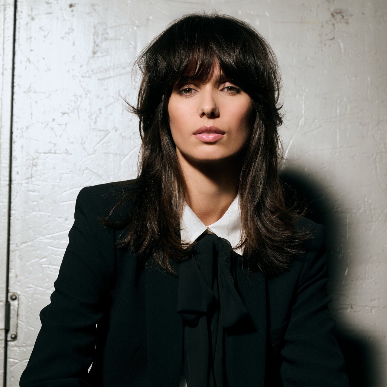 Frau mit dunkelbraunem, stufig geschnittenem Haar und lässigem Pony trägt einen schwarzen Blazer mit Schluppenbluse.