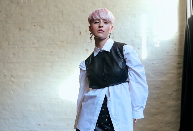 Junge Frau mit kurzem, rosafarbenem Haar posiert selbstbewusst in einem Layering-Look aus weißer Hemdbluse und schwarzer Lederweste vor einer hellen Backsteinwand.