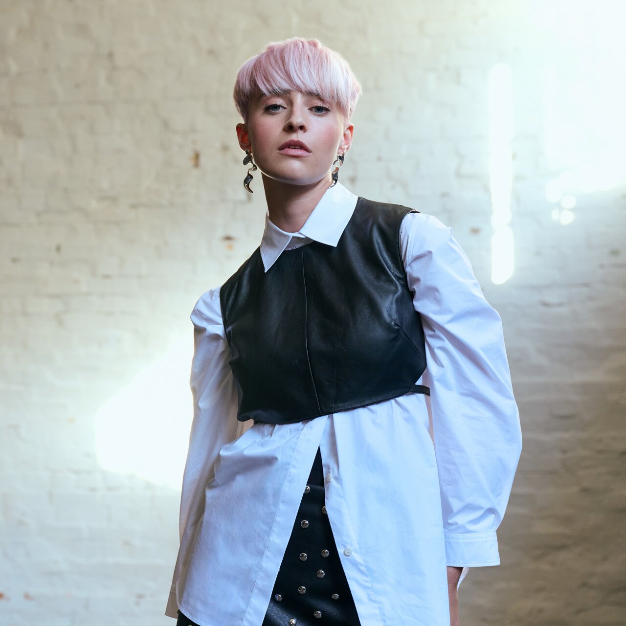 Junge Frau mit kurzem, rosafarbenem Haar posiert selbstbewusst in einem Layering-Look aus weißer Hemdbluse und schwarzer Lederweste vor einer hellen Backsteinwand.