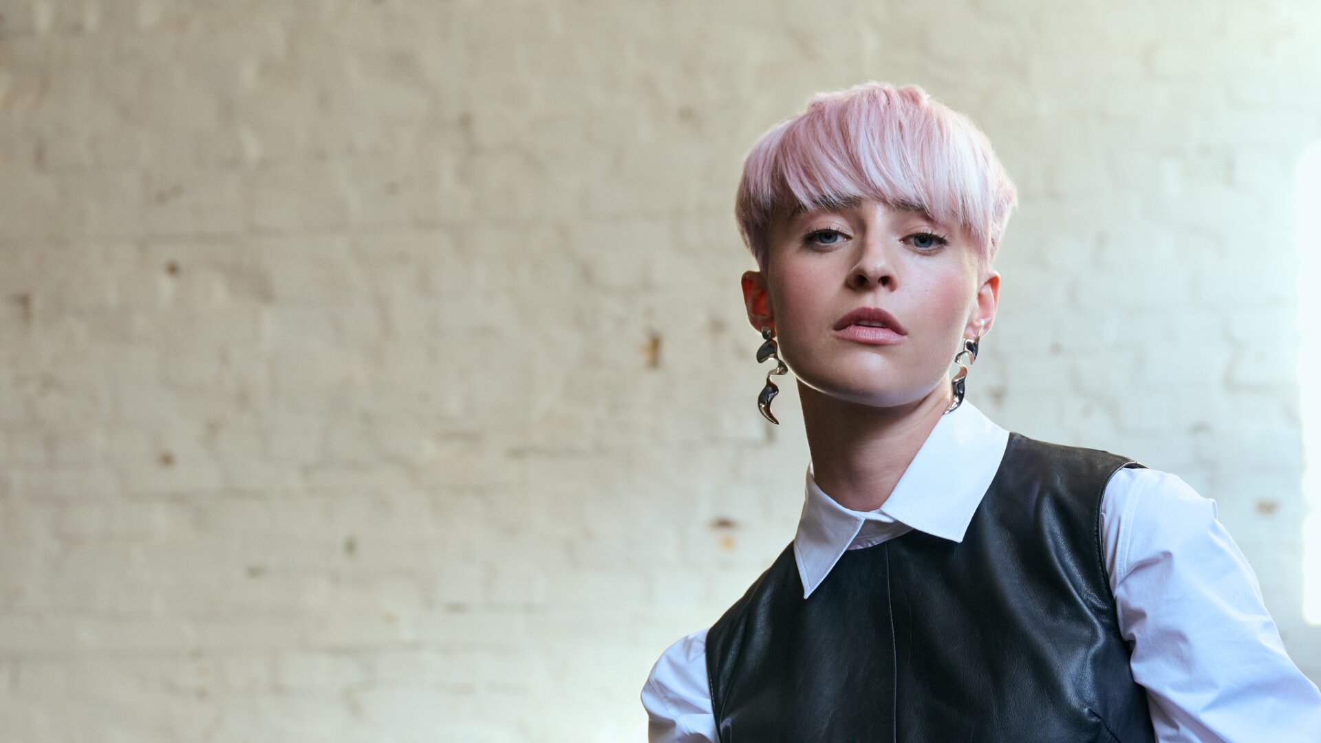 Junge Frau mit kurzem, rosafarbenem Haar posiert selbstbewusst in einem Layering-Look aus weißer Hemdbluse und schwarzer Lederweste vor einer hellen Backsteinwand.