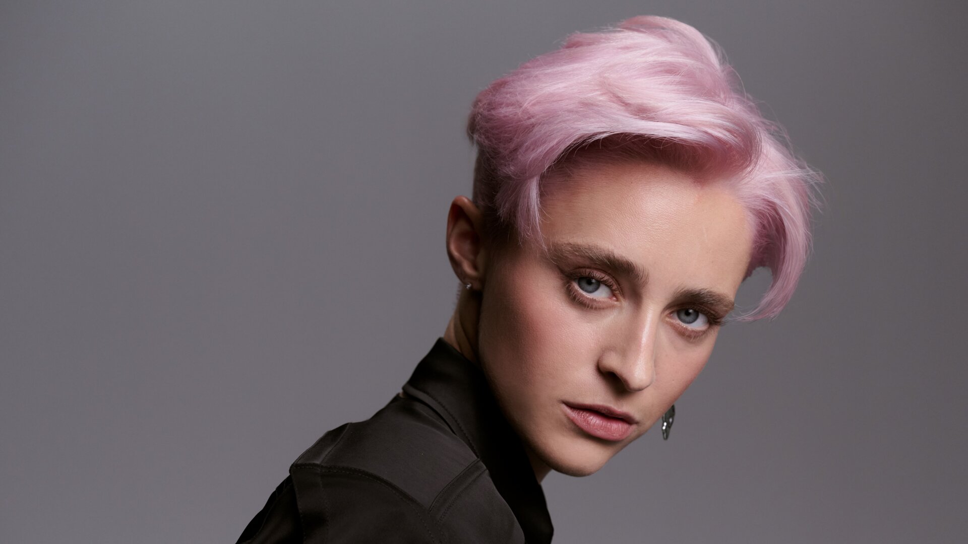 Junge Frau mit kurzem, pastellrosa gefärbtem Haar und natürlichem Make-up, die in einer schwarzen Bluse selbstbewusst über die Schulter blickt.