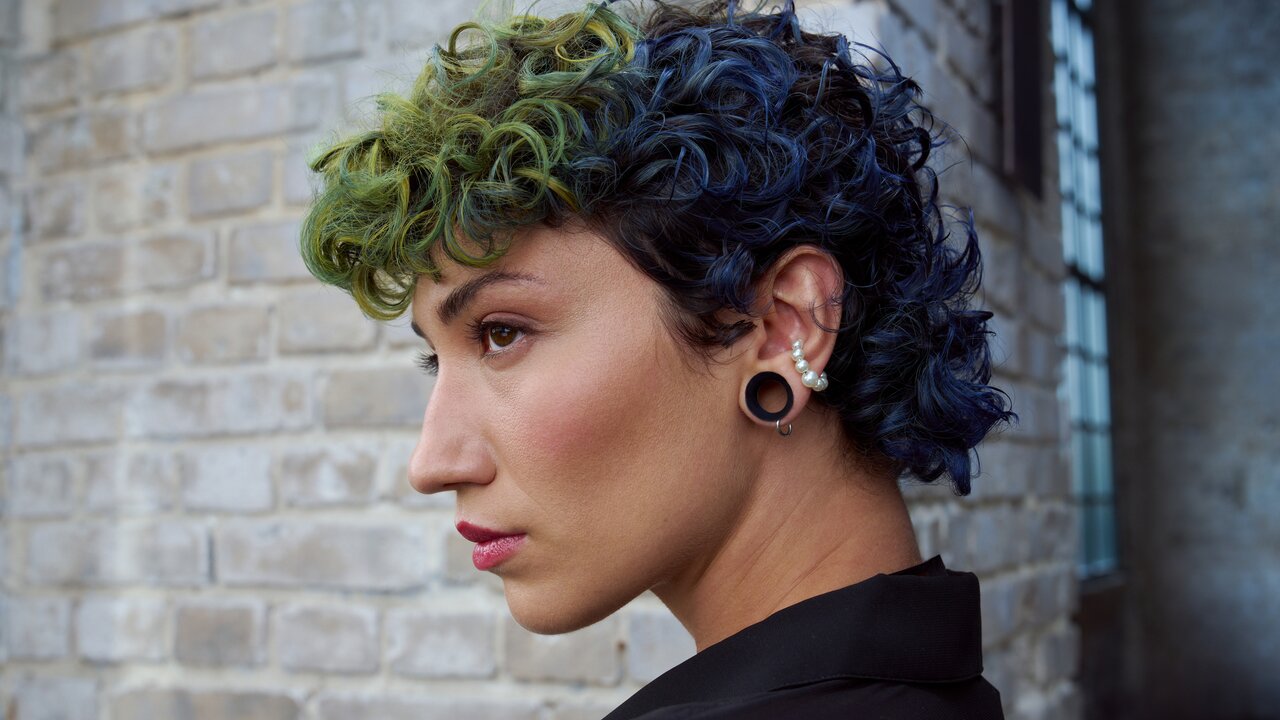 Seitenansicht einer Person mit kurz gelocktem, grün-blauem Haar, mehreren Ohrpiercings und schwarzer Kleidung vor einer Backsteinwand.