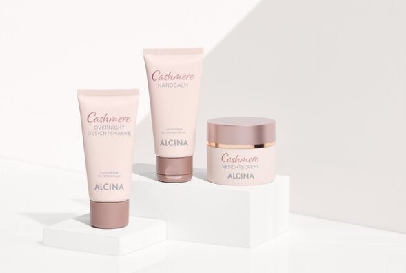 ALCINA Cashmere Pflegeserie mit Overnight Gesichtsmaske, Handbalm und Gesichtscreme in zart roséfarbenem Design, stilvoll auf weißen Podesten inszeniert.