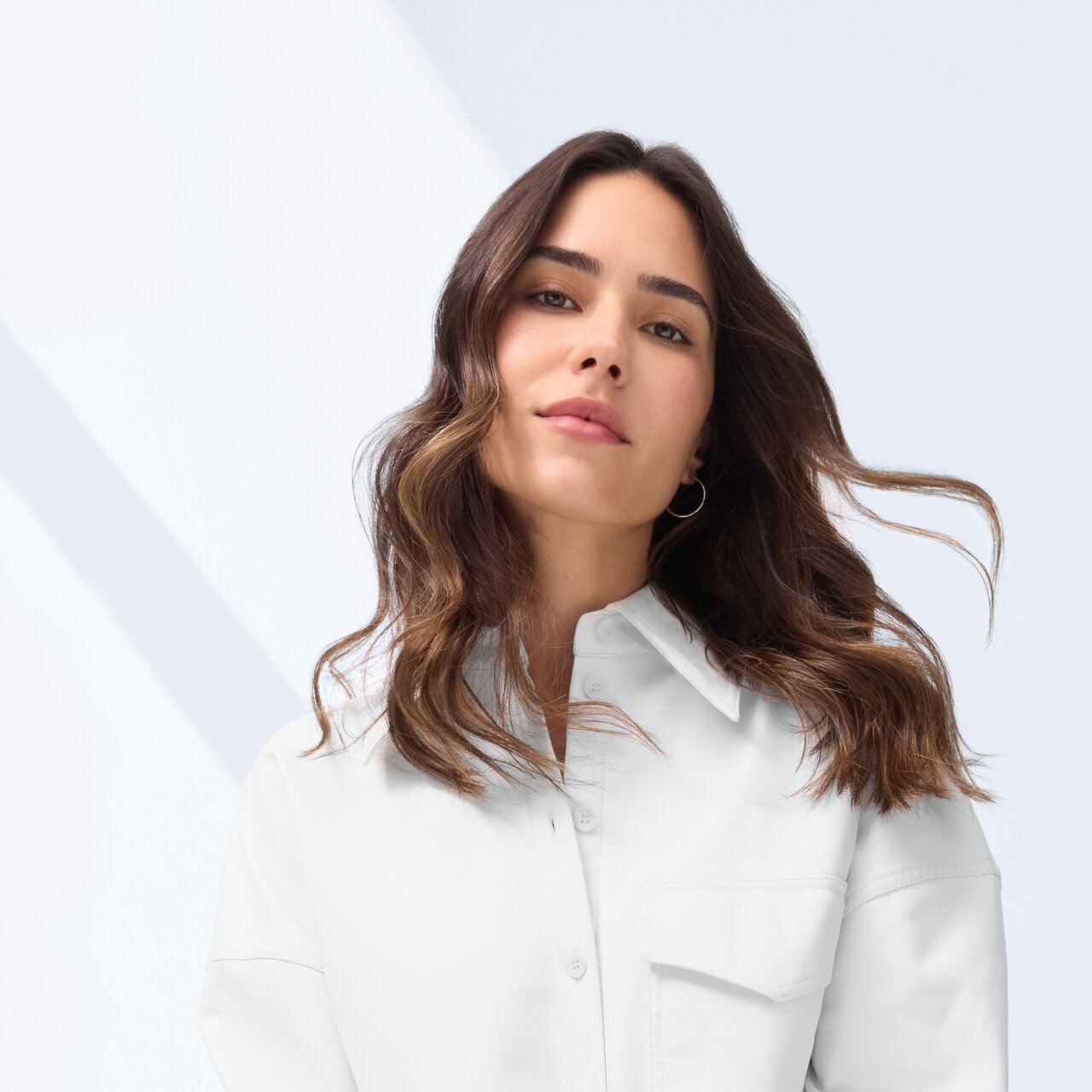 Frau mit langem, welligem dunkelbraunem Haar sitzt entspannt in einer weißen Oversize-Hemdjacke und einer dunkelgrauen weiten Hose.