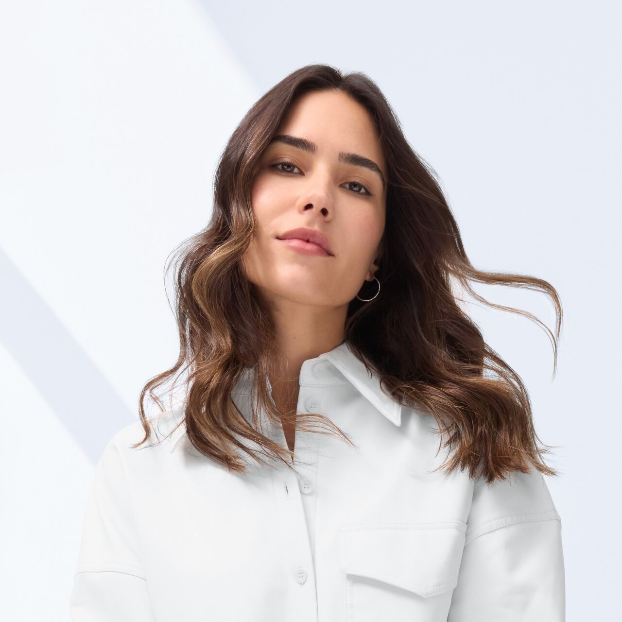 Frau mit langem, welligem dunkelbraunem Haar sitzt entspannt in einer weißen Oversize-Hemdjacke und einer dunkelgrauen weiten Hose.