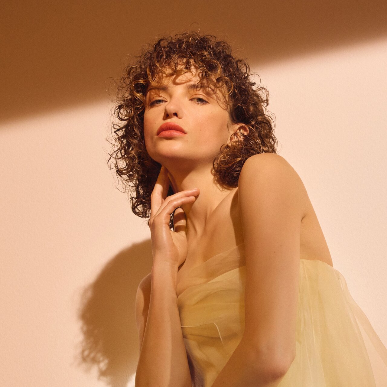 Junge Frau mit natürlichen, definierten Locken und kurzem Pony posiert in einem zarten, hellgelben Tüllkleid im warmen Sonnenlicht.