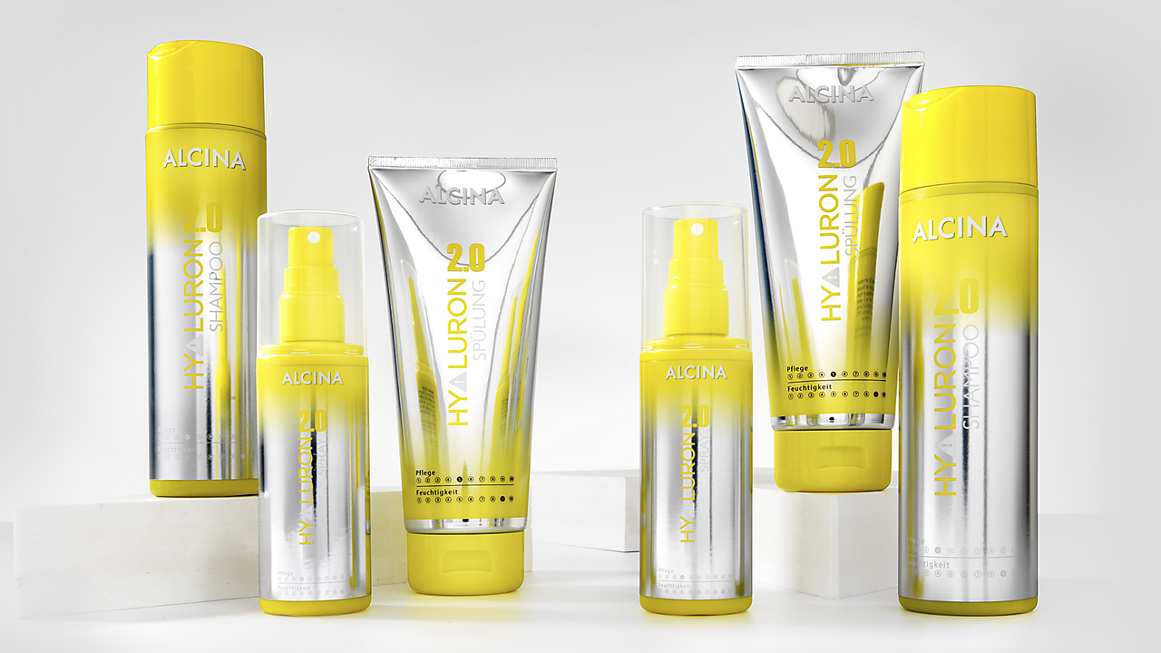 ALCINA Hyaluron-Pflegeserie in gelb-silbernem Design, bestehend aus Shampoo, Spülung und Styling-Produkten, die nebeneinander auf weißen Podesten für eine moderne Haarpflegepräsentation arrangiert sind.