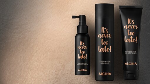 ALCINA Its never too late Coffein Vital Pflegeserie mit Kopfhaut-Serum, Shampoo und Conditioner in mattschwarzen Verpackungen mit kupferfarbener Schrift, stilvoll auf einem warm beleuchteten Hintergrund arrangiert.
