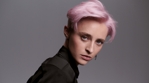 Junge Frau mit kurzem, pastellrosa gefärbtem Haar und natürlichem Make-up, die in einer schwarzen Bluse selbstbewusst über die Schulter blickt.