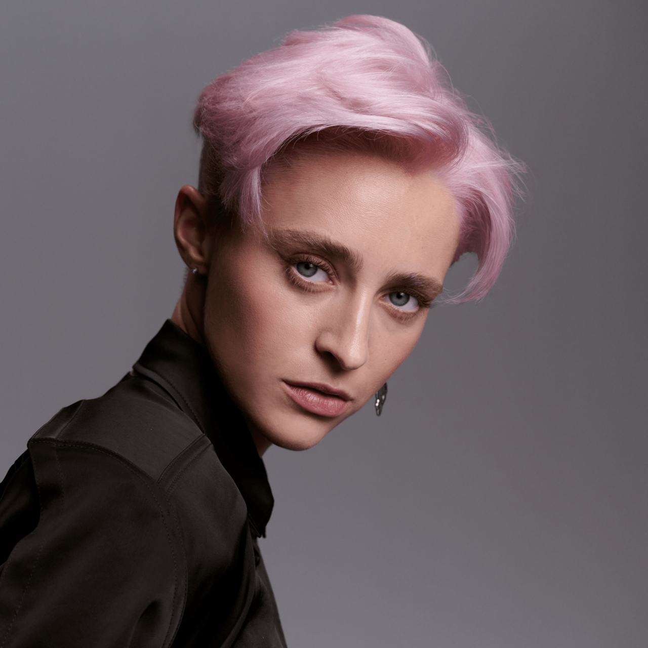 Junge Frau mit kurzem, pastellrosa gefärbtem Haar und natürlichem Make-up, die in einer schwarzen Bluse selbstbewusst über die Schulter blickt.