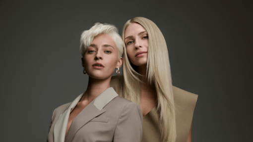 Zwei Frauen in eleganten beigefarbenen Outfits posieren nebeneinander – eine mit kurzem, platinblondem Haar und eine mit langem, warmen Blondhaar.