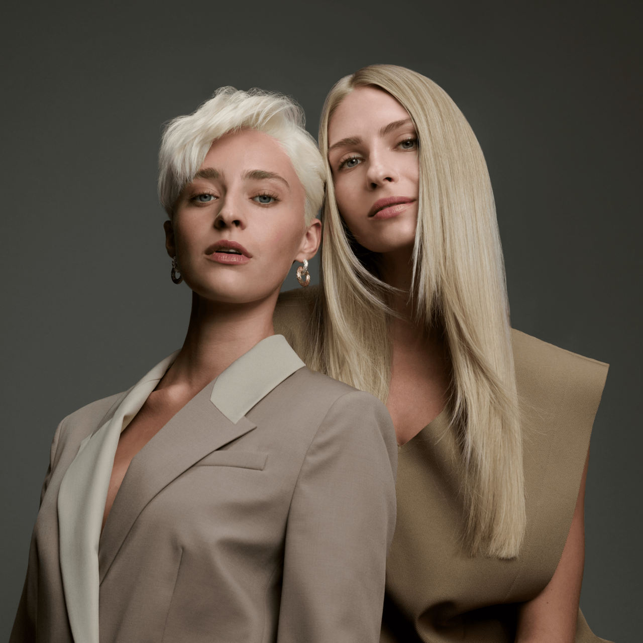 Zwei Frauen in eleganten beigefarbenen Outfits posieren nebeneinander – eine mit kurzem, platinblondem Haar und eine mit langem, warmen Blondhaar.