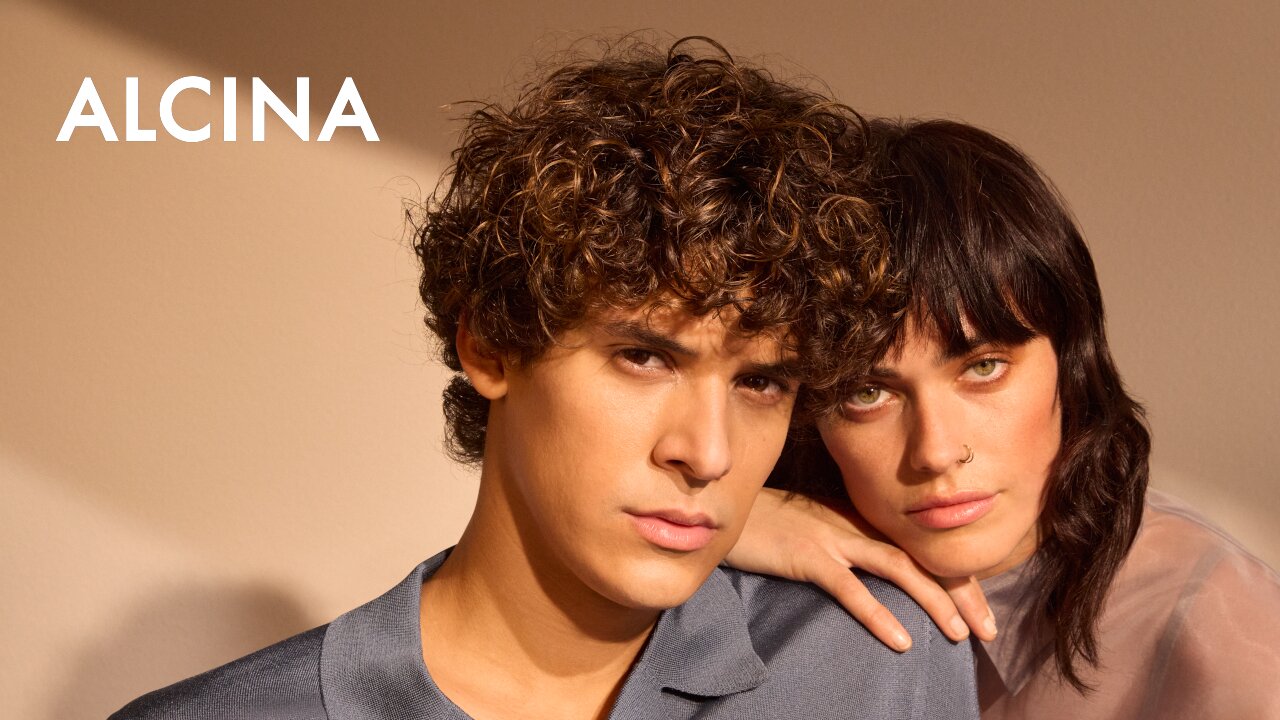 ALCINA Kampagnenmotiv mit einem jungen Mann mit voluminösen braunen Locken und einer Frau mit dunklem, welligem Haar und Pony, die sich vertraut aneinander lehnen und natürliche Haarstylings präsentieren.